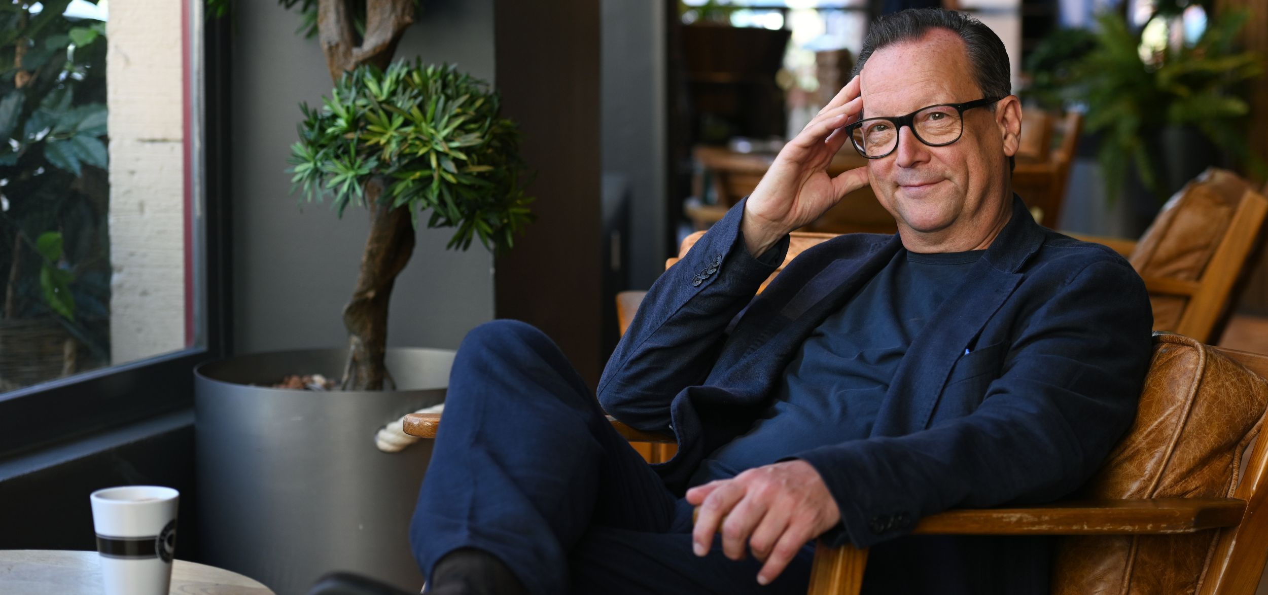 Schauspieler Matthias Brandt in dunkelblauem Blazer, entspannte Sitzposition vor Zimmerpflanzen