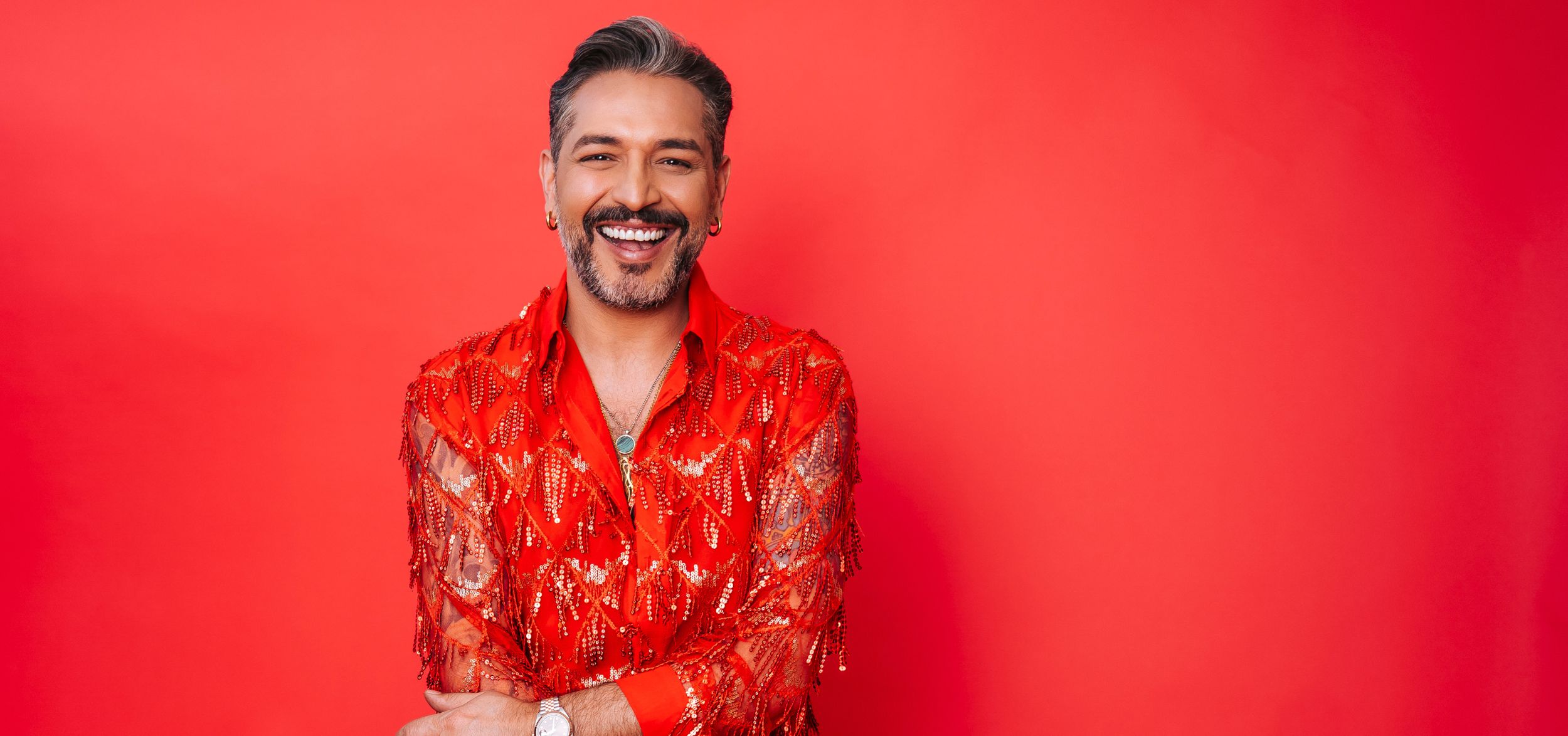 Schauspieler Gianni Jovanovic in rotem Pailletten-Jacket, breit lächelnd vor rotem Hintergrund