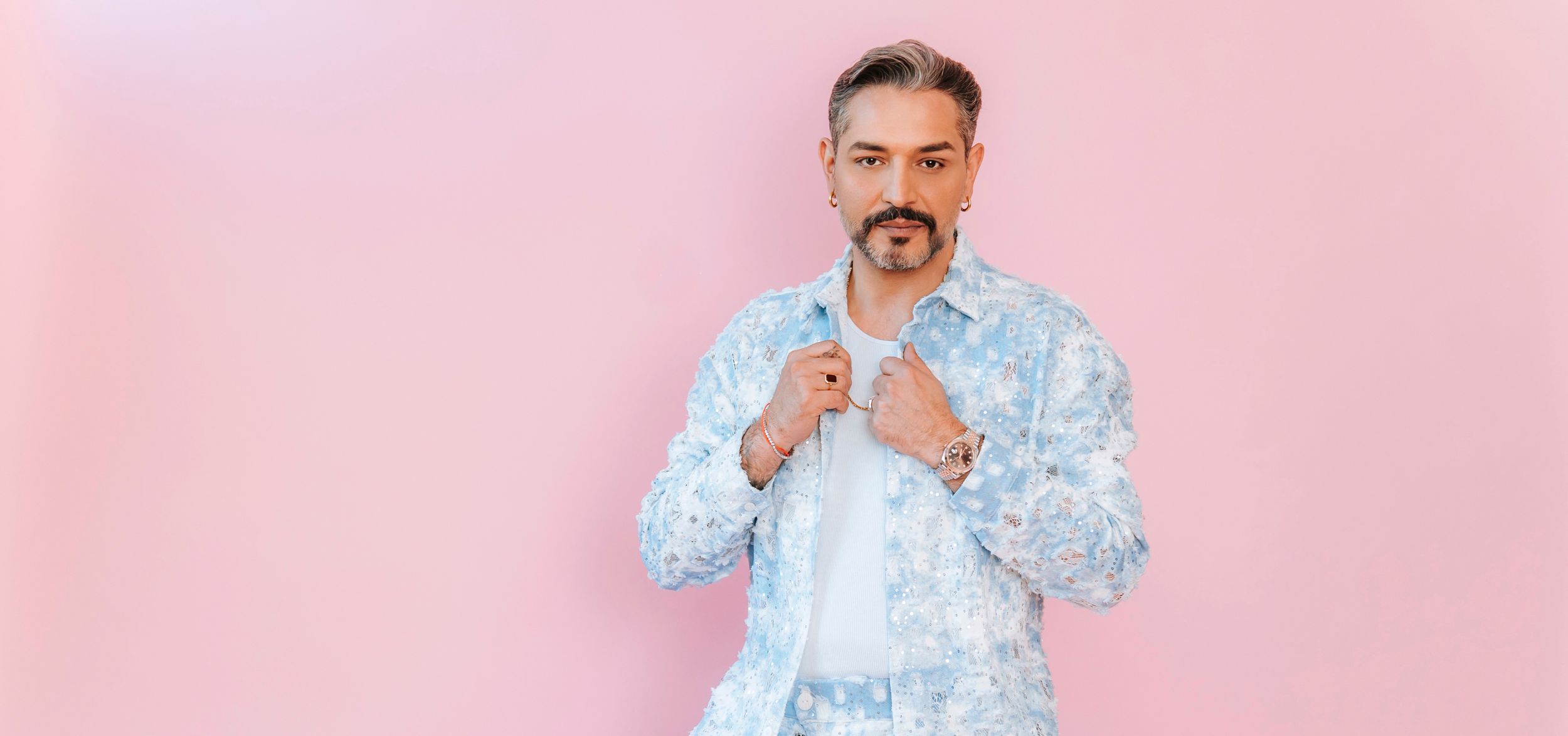 Schauspieler Gianni Jovanovic in hellblauem Blumen-Jackett, nachdenkliche Geste vor rosafarbenem Hintergrund
